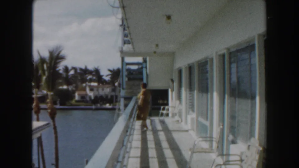 Woman continues along the waterfront balcony in 1960 Miami footage, passing windows and chairs with canal water at left.