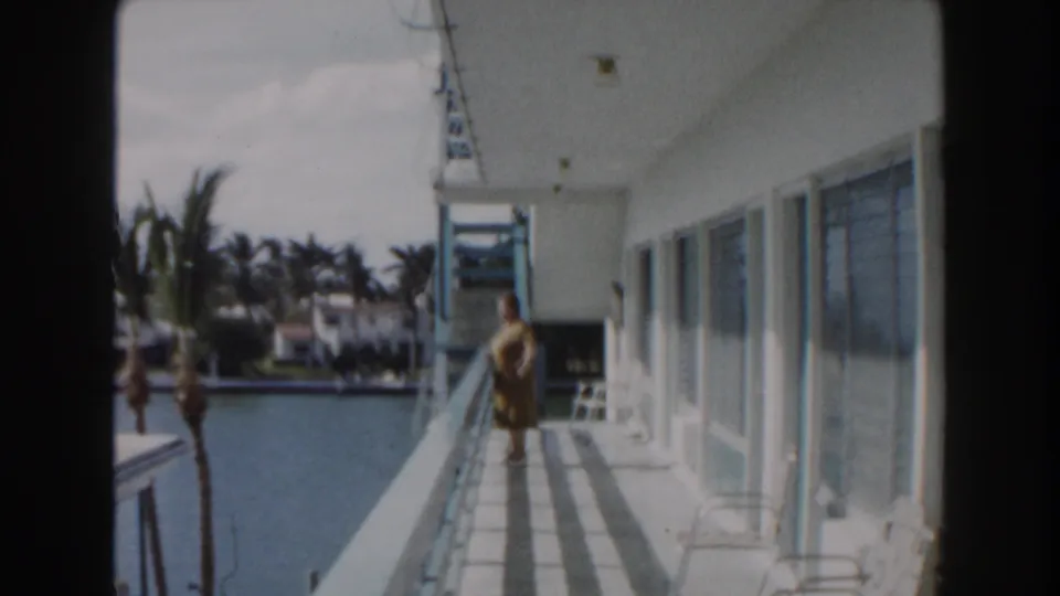 Woman in a yellow dress stands or walks on a Miami balcony, with canal water, palms, and waterfront houses visible beyond.