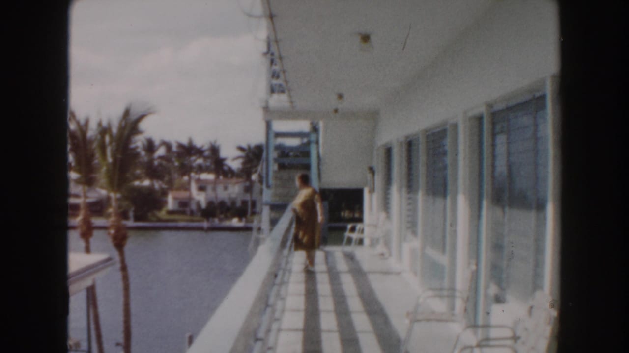 Elderly woman walking on a balcony beside a canal with palm trees and white buildings nearby.