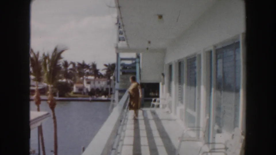 1960 Miami home-movie frame of an elderly woman in a yellow dress walking along a sunlit balcony overlooking a canal.