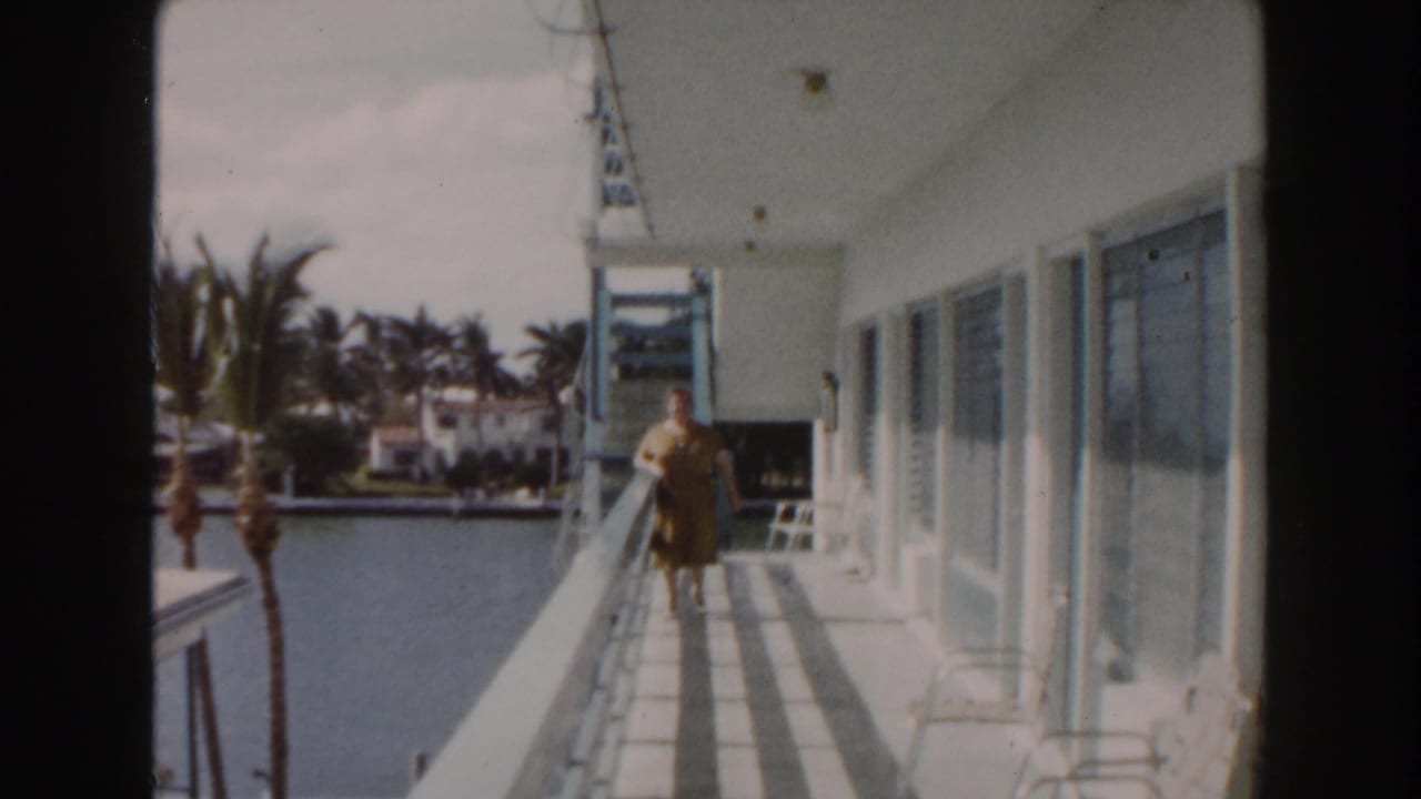 Woman standing farther along a balcony above canal water with palm trees across the way.