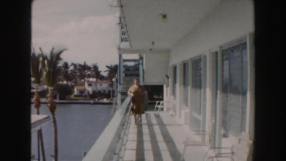 1960 Miami balcony view with an elderly woman near the railing, canal water to the left, and white waterfront buildings beyond.