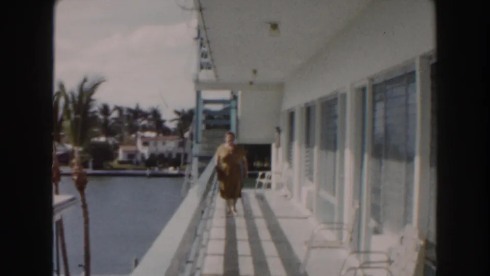 1960 Miami home-movie frame showing an elderly woman approaching the camera along a balcony with waterfront scenery at left.