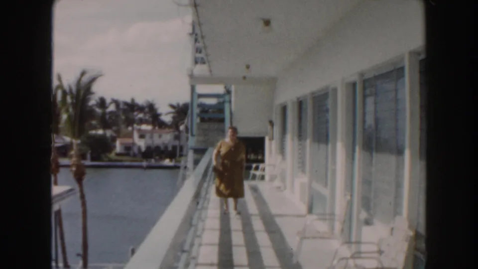 1960 Miami frame of an elderly woman near the camera on a waterfront balcony, with canal water and palm trees behind her.