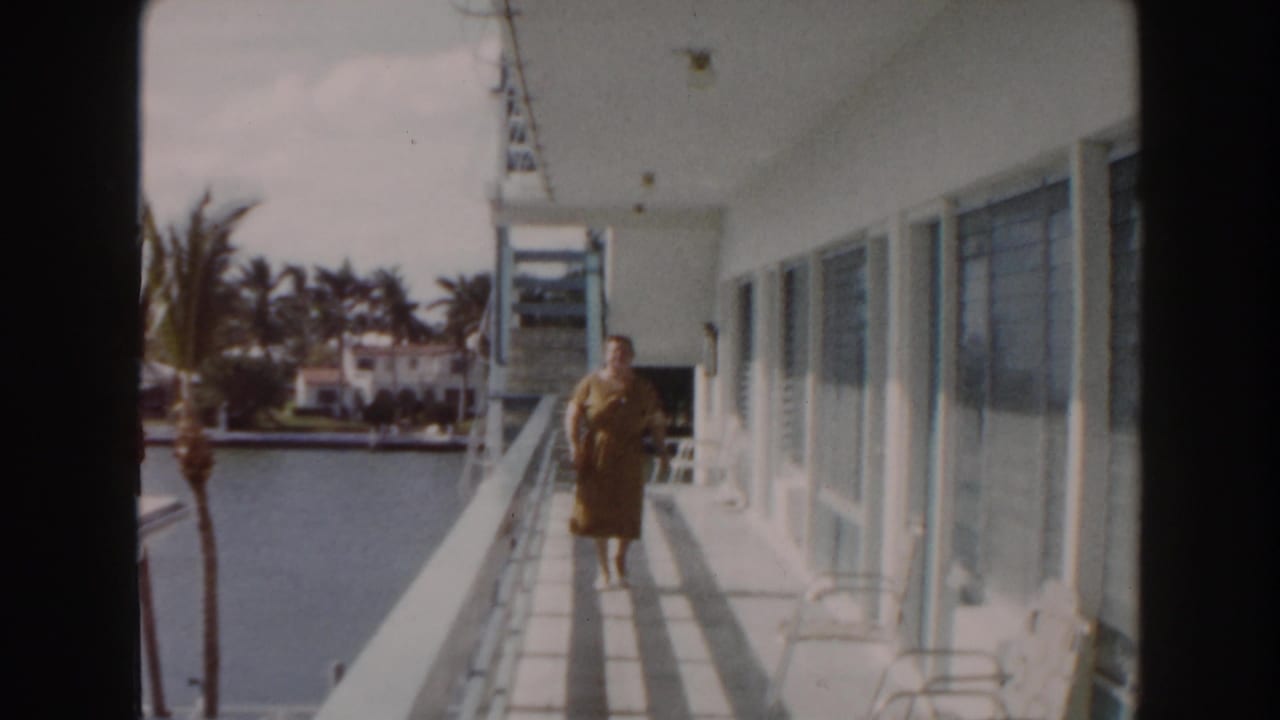 In 1960 Miami home-movie footage, a woman in a brown dress walks on a sunlit balcony overlooking water and palm trees.