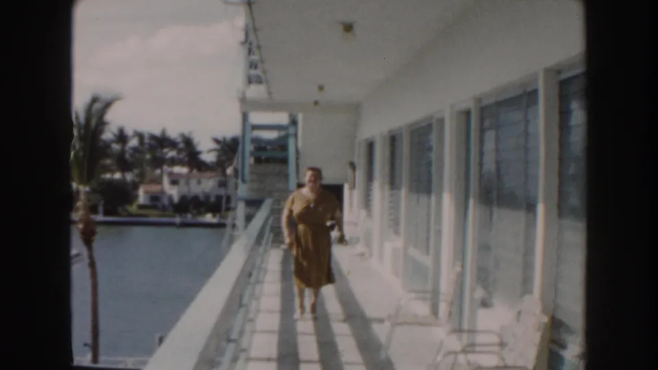 A woman in a brown dress approaches the camera along a Miami waterfront balcony, with white walls, windows, water, and palms visible.