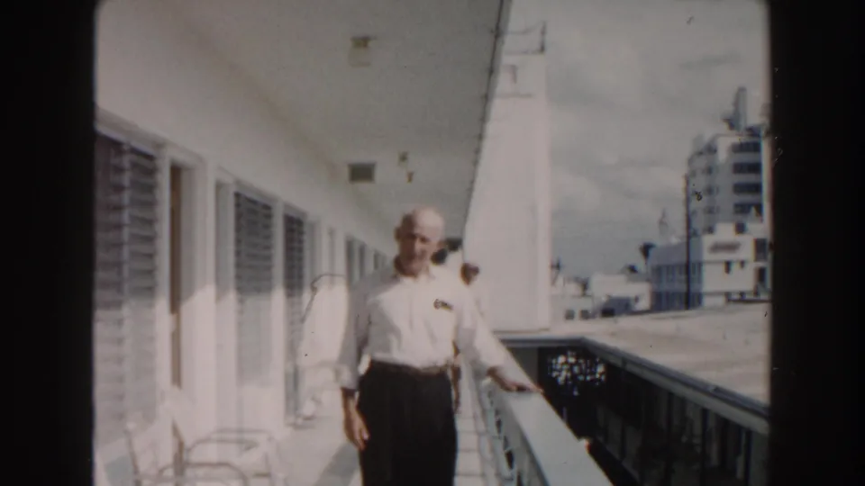 An older man in a white shirt stands on a Miami balcony, with louvered windows, railings, and nearby city buildings behind him.