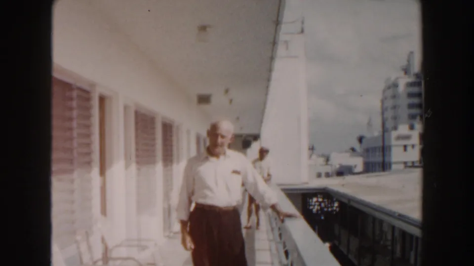 An older man stands near a balcony railing in 1960 Miami footage, with a partly visible person, white exterior walls, and buildings beyond.