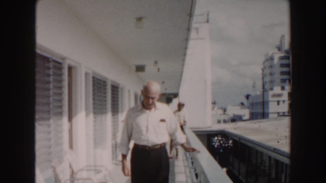 In 1960 Miami home-movie footage, an older man in a white shirt walks toward the camera along an outdoor hotel corridor.