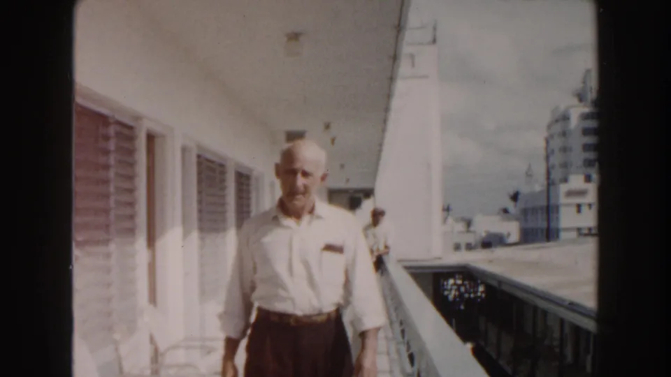The man continues down the Miami outdoor hotel walkway, with columns, railing, and mid-century buildings visible in the background.
