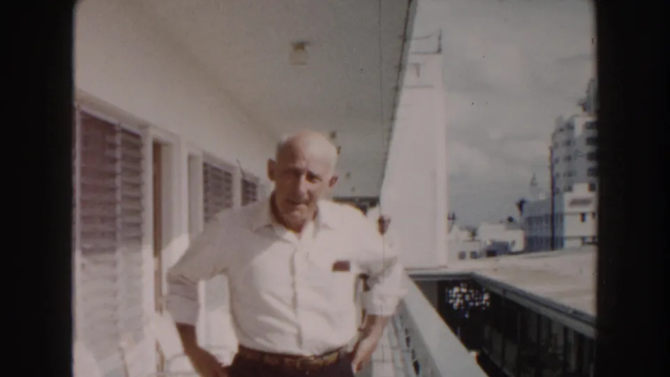 A close view shows the older man pausing or slowing on the sunlit Miami hotel corridor, with louvered windows along the wall.