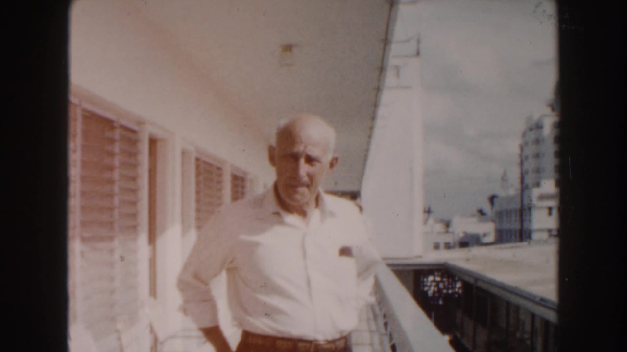 In 1960 Miami home-movie footage, an elderly bald man in a white shirt stands on a sunlit balcony near louvered windows and distant buildings.