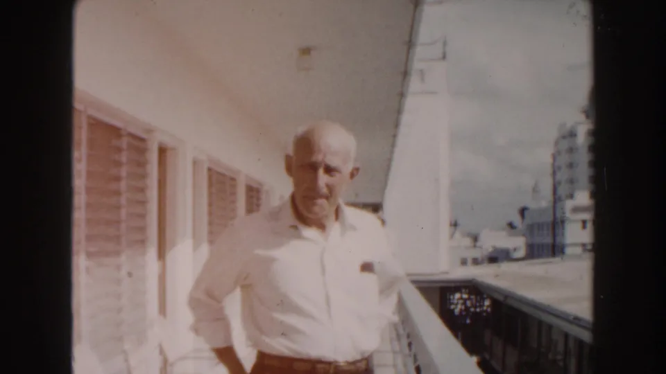 In 1960 Miami home-movie footage, an elderly bald man in a white shirt stands on a sunlit balcony near louvered windows and distant buildings.