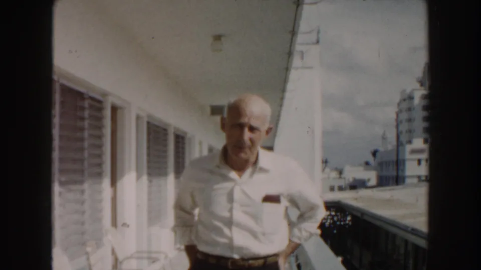 A softly focused 8mm frame shows the elderly man on the Miami balcony, partly turned under the overhang with distant buildings beyond the rail.