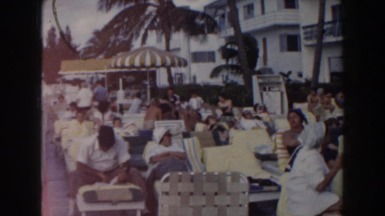 1960 Miami home-movie frame of guests seated and reclining poolside near a striped umbrella, palm trees, and white buildings.