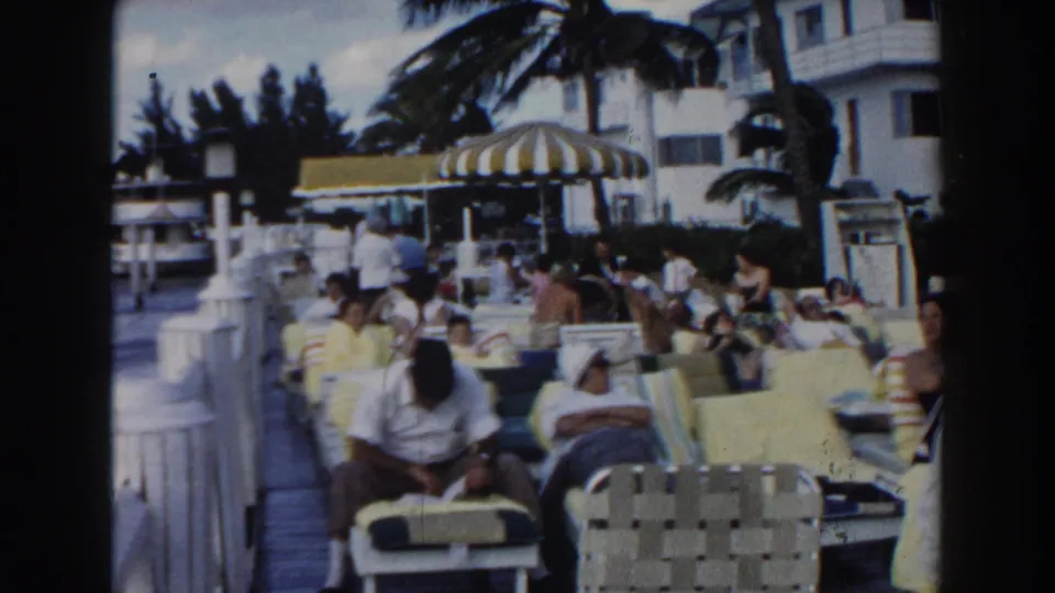 1960 Miami resort pool scene with people relaxing on lounge chairs beneath a striped umbrella and palm trees.