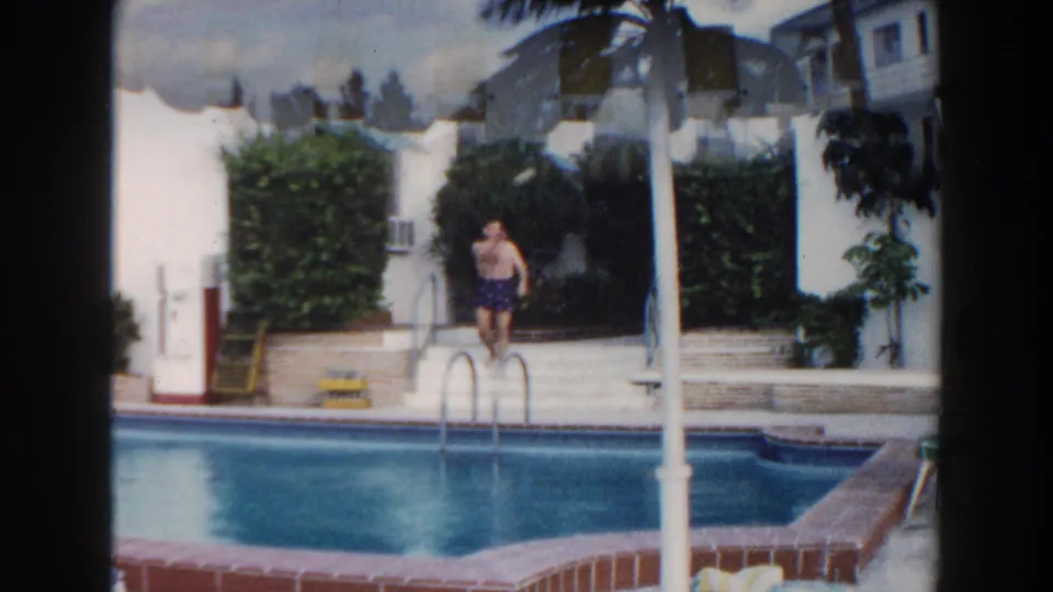 1960 Miami pool frame showing a man near the ladder, blue water, striped shade, hedges, and white resort walls.