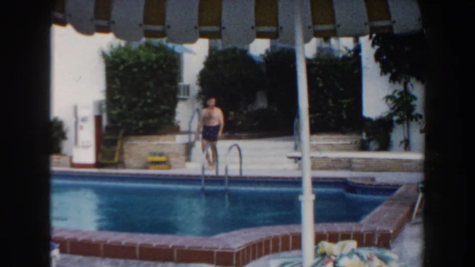 1960 Miami home-movie frame of a man at the pool edge, with striped shade, hedges, white walls, and blue water visible.
