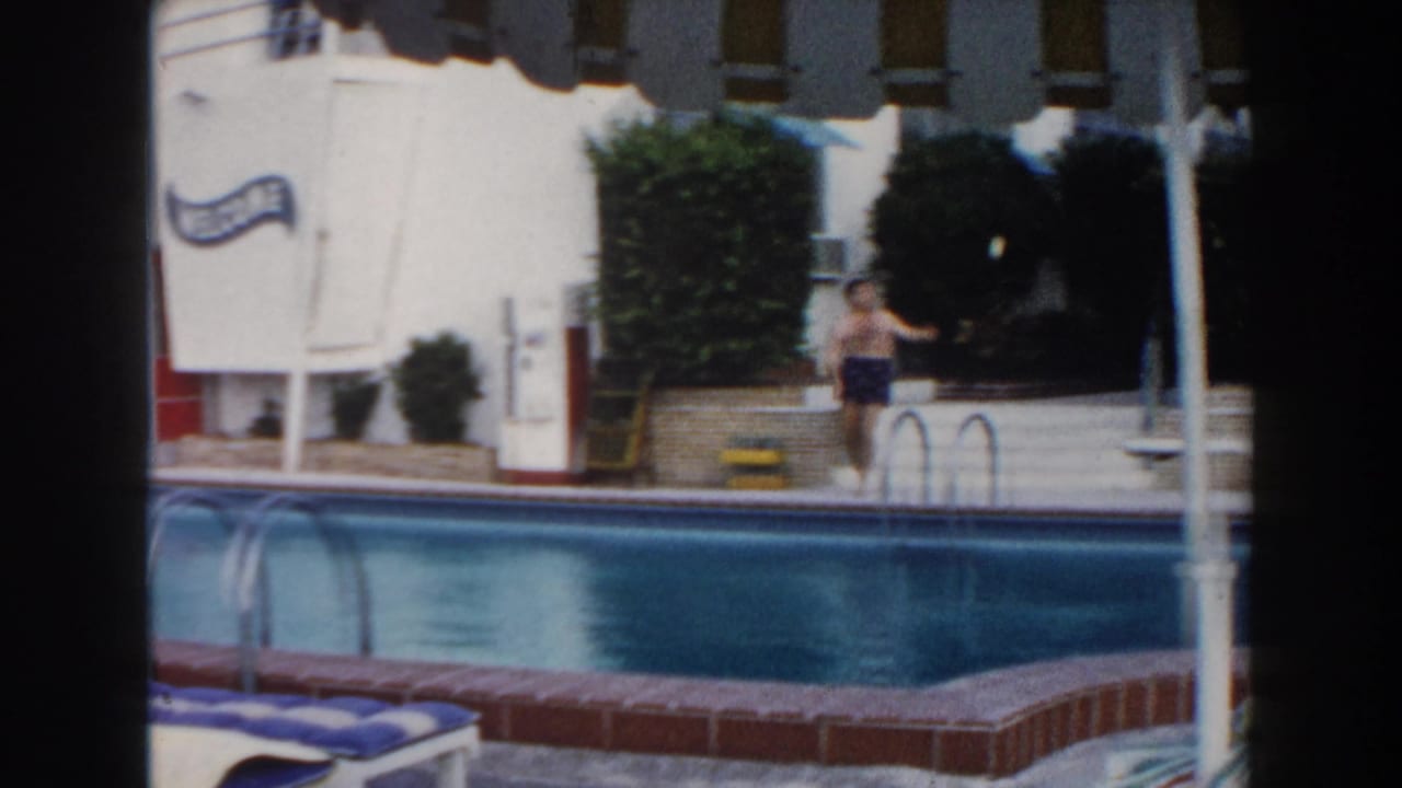 1960 Miami home-movie frame showing a man by a motel pool, blue loungers, white building walls, shrubs, and a WELCOME sign.
