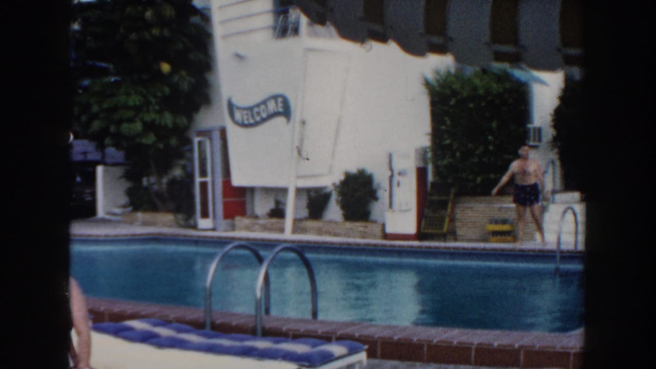 Man walks near the pool edge in front of a white motel wall and WELCOME sign.