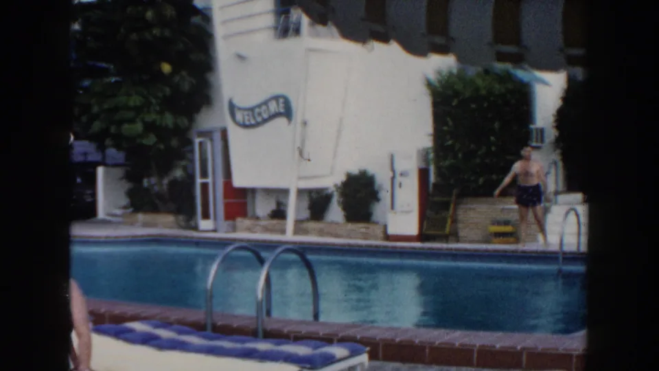 1960 Miami poolside frame with a man in dark swim trunks near the ladder, white motel facade, shrubs, and visible WELCOME sign.