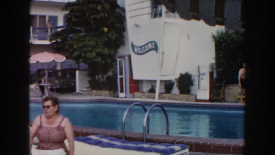 1960 Miami home-movie frame showing a seated woman by a motel pool with striped awning, pool ladder, shrubs, and WELCOME sign.
