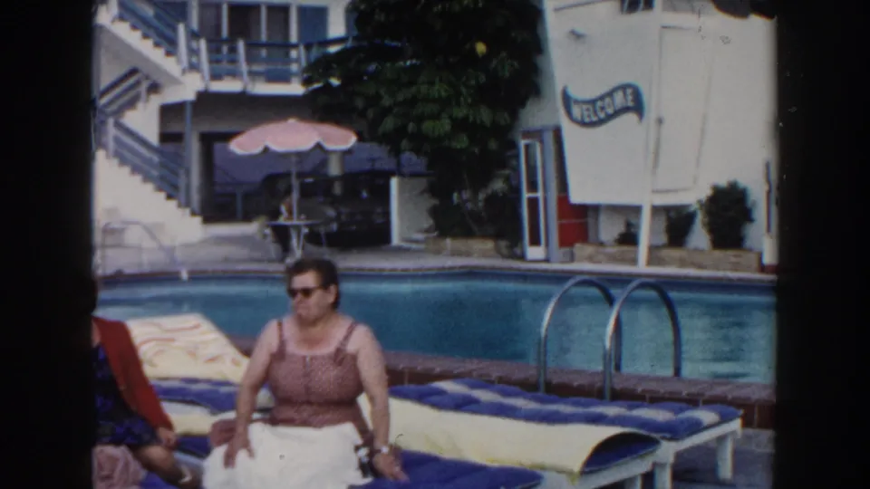1960 Miami motel pool frame with a woman seated on a blue-cushioned lounger, another guest reclining, pool ladder, stairs, and awning.