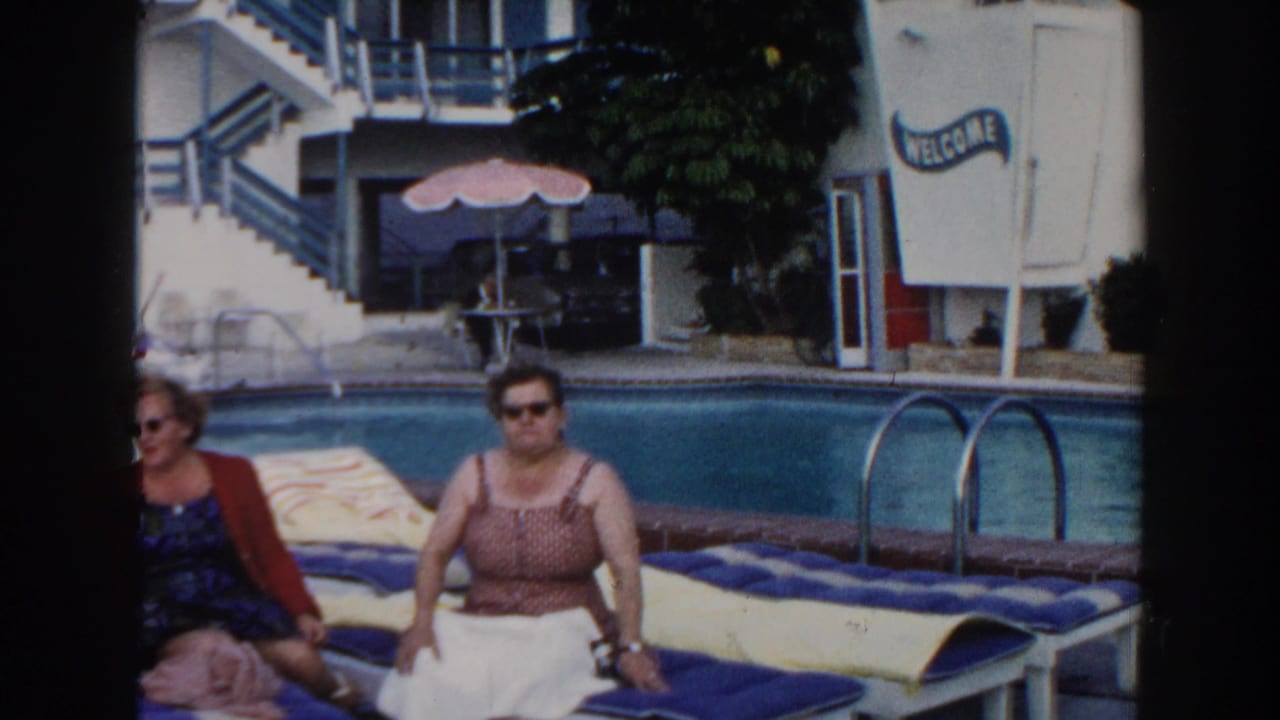 Two adult women sit on poolside lounge chairs in Miami, Florida, with a white resort building and WELCOME sign behind them.