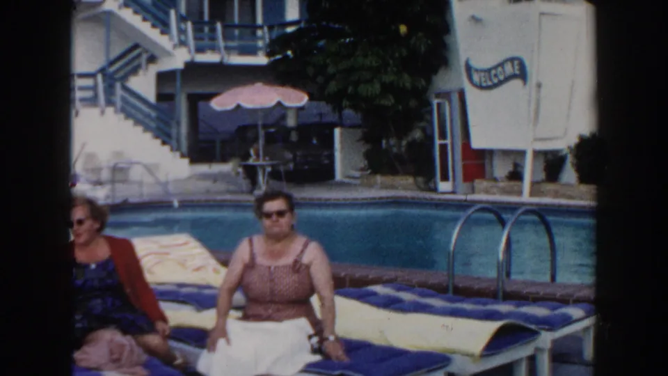 Two adult women sit on poolside lounge chairs in Miami, Florida, with a white resort building and WELCOME sign behind them.