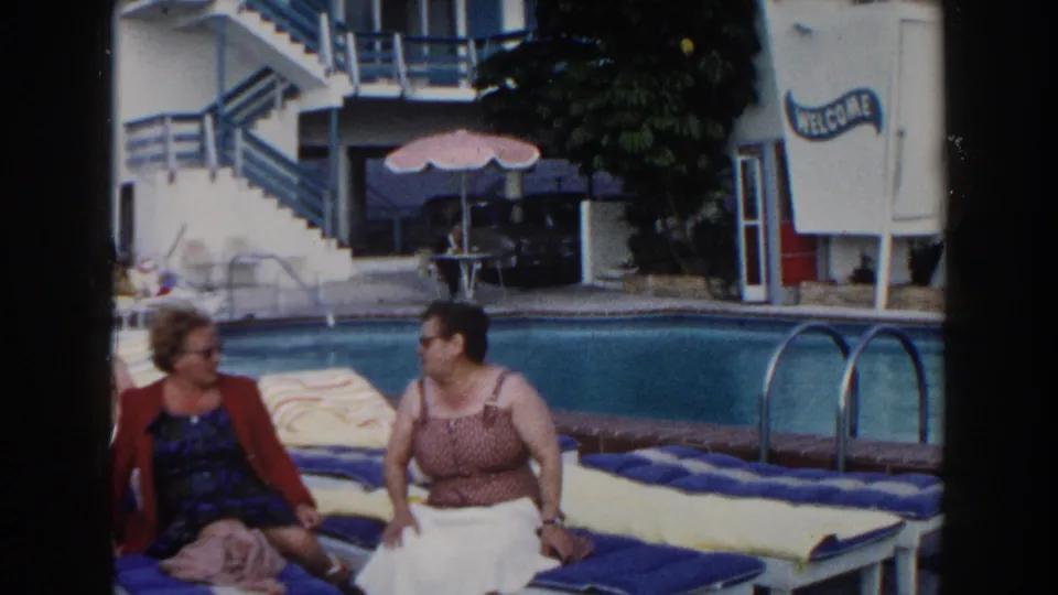 Two adult women sit beside a Miami resort pool, one turning toward the other during a casual conversation.