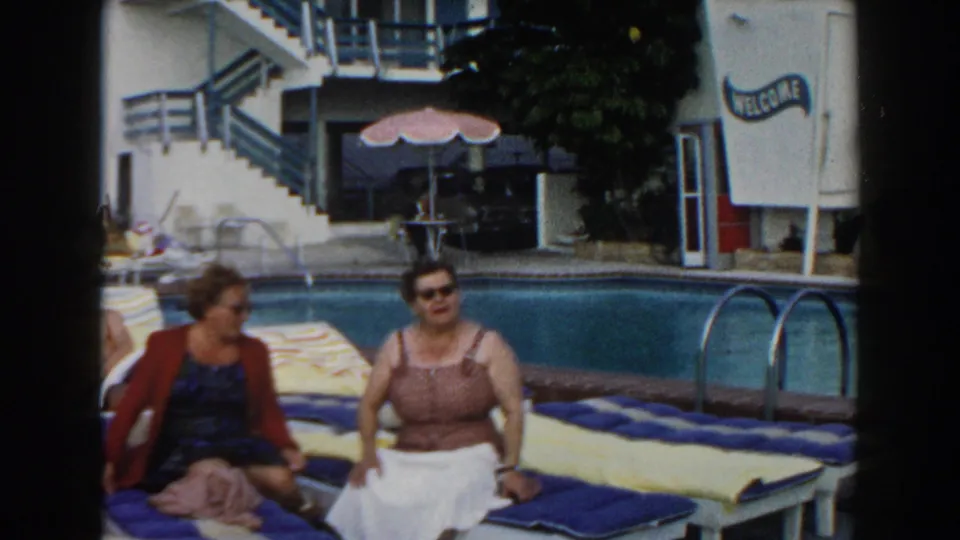 Two adult women remain seated on cushioned lounge chairs by a Miami resort swimming pool, with stairs and a WELCOME sign behind them.
