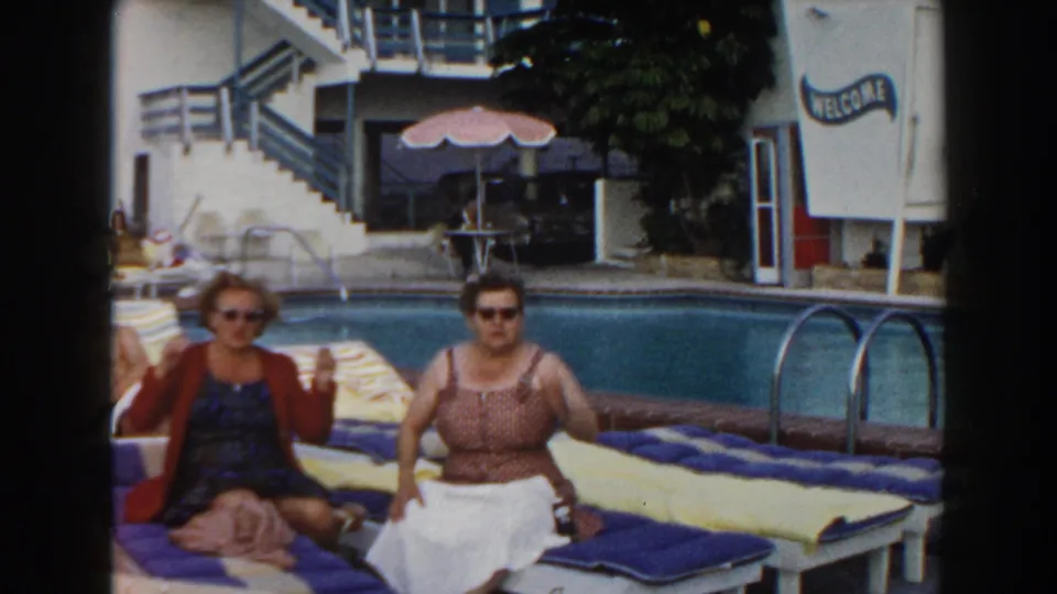 Two adult women gesture from lounge chairs beside a Miami resort pool, with a pink umbrella and WELCOME sign visible behind them.