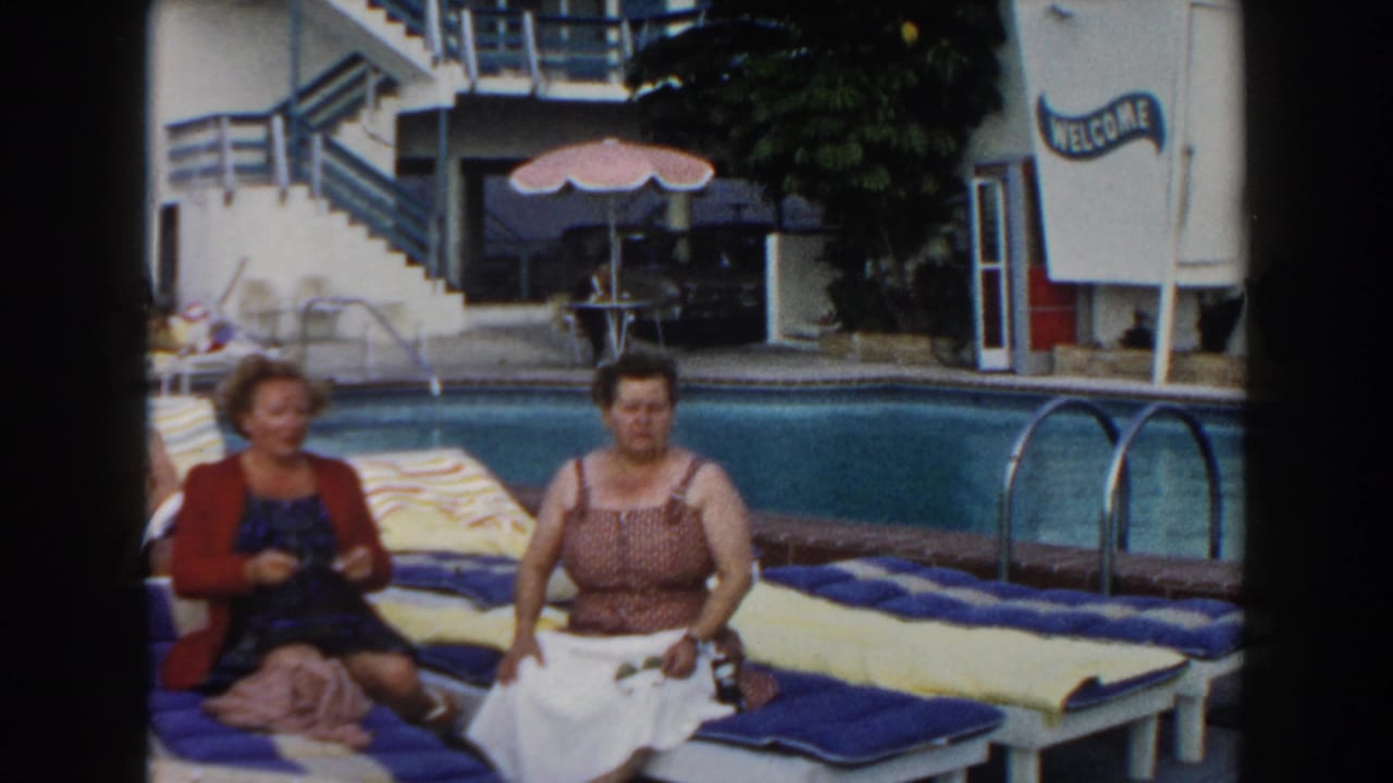 1960 Miami poolside home-movie frame showing two women seated on blue and yellow lounge chairs near a pool and motel-style stairs.