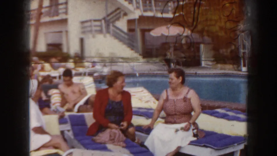 1960 Miami poolside frame with two women conversing on striped lounge chairs as sunbathers and a white building appear behind them.