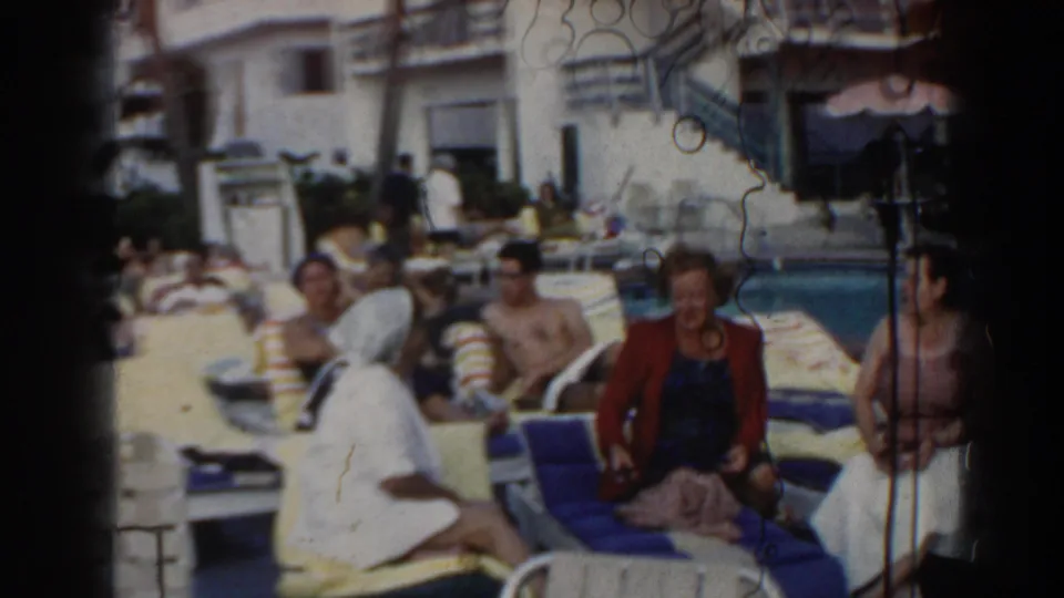 1960 Miami home-movie frame showing several adults around a pool, including women seated on lounge chairs and sunbathers in the background.