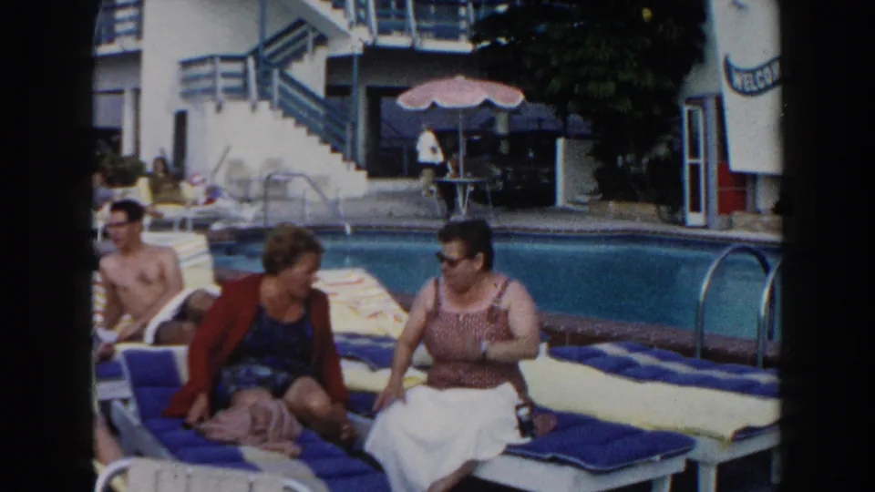 1960 Miami poolside frame showing two women on blue and yellow lounge chairs near the pool, with guests and motel-style railings behind them.