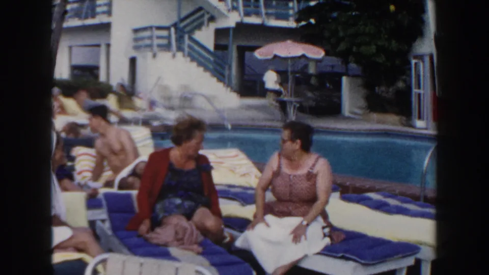 In supplied 1960 Miami footage, two women relax and talk on blue loungers by a hotel pool with guests and a stairway behind them.