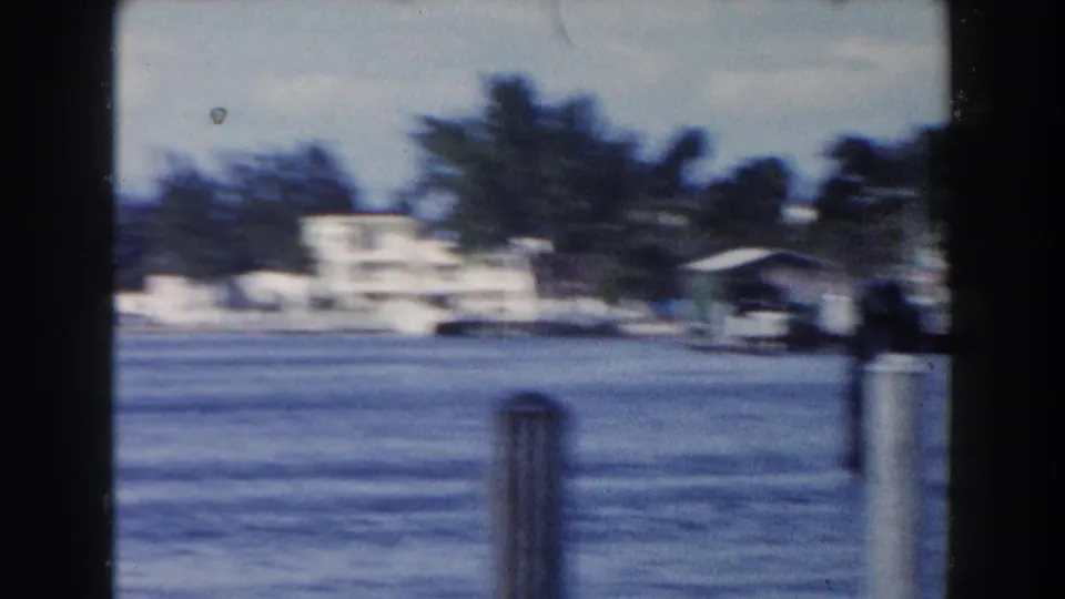 A handheld 8mm view looks across Miami water toward dock posts, palm trees, and low waterfront buildings in soft focus.