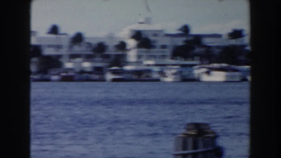 1960 Miami home-movie frame with motion blur over blue water, white shoreline buildings, palm trees, and docked boats.