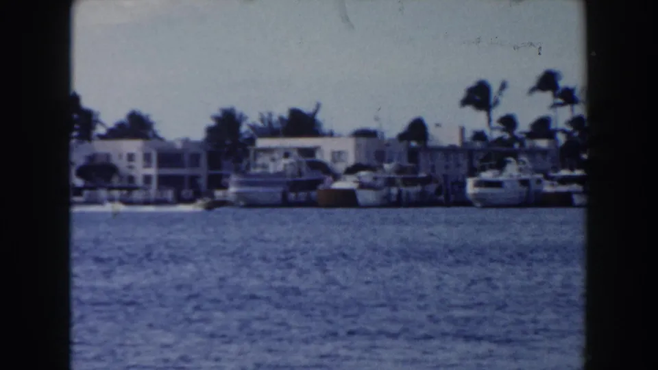 1960 Miami waterfront frame showing white low-rise buildings, palm trees, moored boats, and rippled blue water.