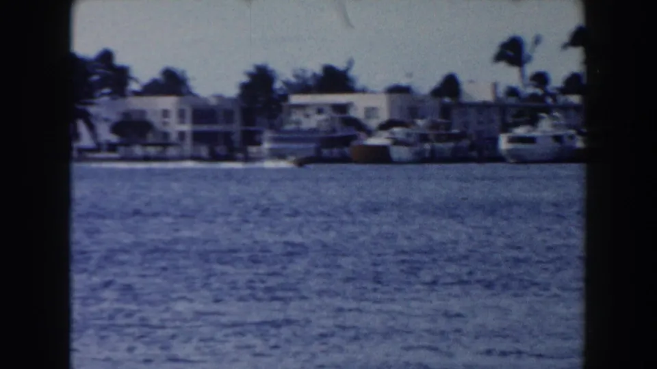 1960 Miami waterfront view across blue water toward low buildings, boats, and palm trees in soft 8mm color.