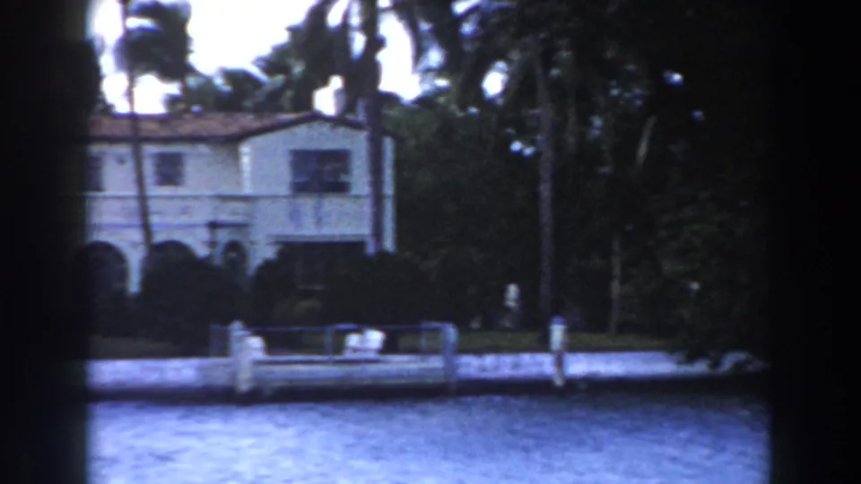 1960 Miami waterfront residence with arched lower openings, palms, shrubs, and a low dock or seawall along the water.