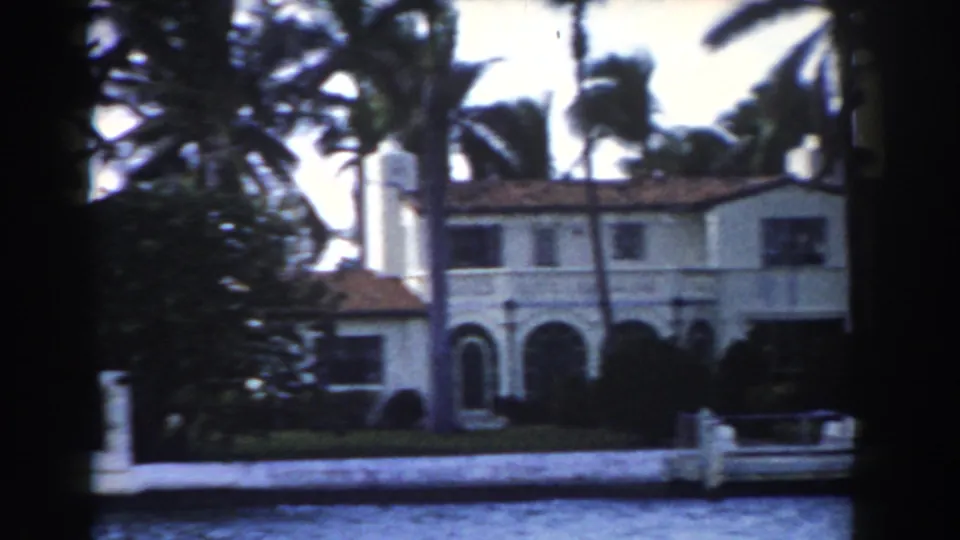 1960 Miami waterfront house with palms, a chimney, arched entry details, and a low wall along the waterline.
