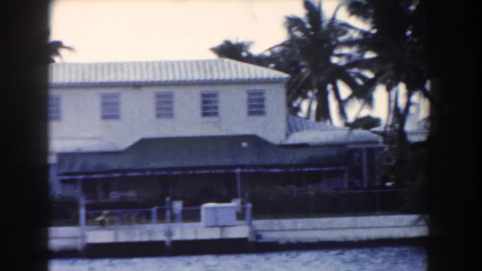1960 Miami frame of a white two-story waterfront building with palms, a dark awning, railings, and water in the foreground.