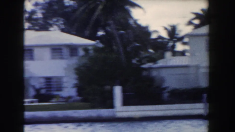 1960 Miami home-movie view of a white low-rise building behind palms and shrubs, with lawn, railings, and water along the foreground.