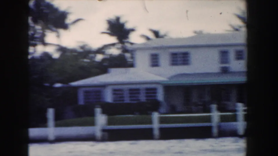 1960 Miami frame showing a white residential-style waterfront building with a green awning, palm trees, lawn, fence posts, and water.