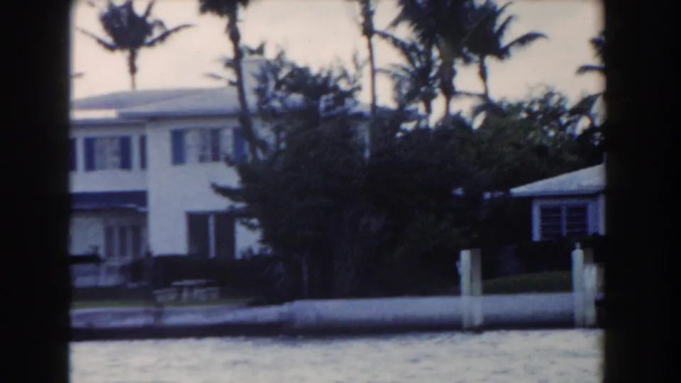 1960 Miami home-movie frame showing white waterfront houses, palms, and a low wall viewed from a boat or shoreline.