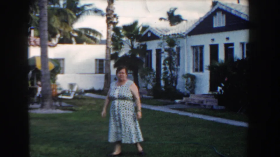 Adult woman walks across a Miami lawn in 1960 home-movie footage, with patio seating, palms, and a white house behind her.