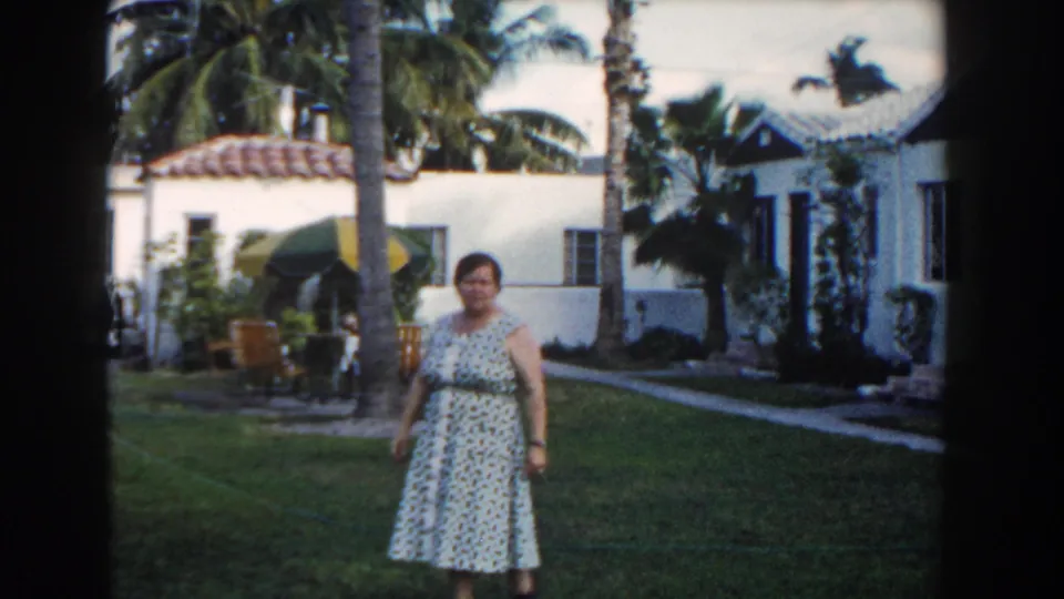 1960 Miami home-movie frame of a woman on a residential lawn with palms, an umbrella, patio seating, and white waterfront houses.