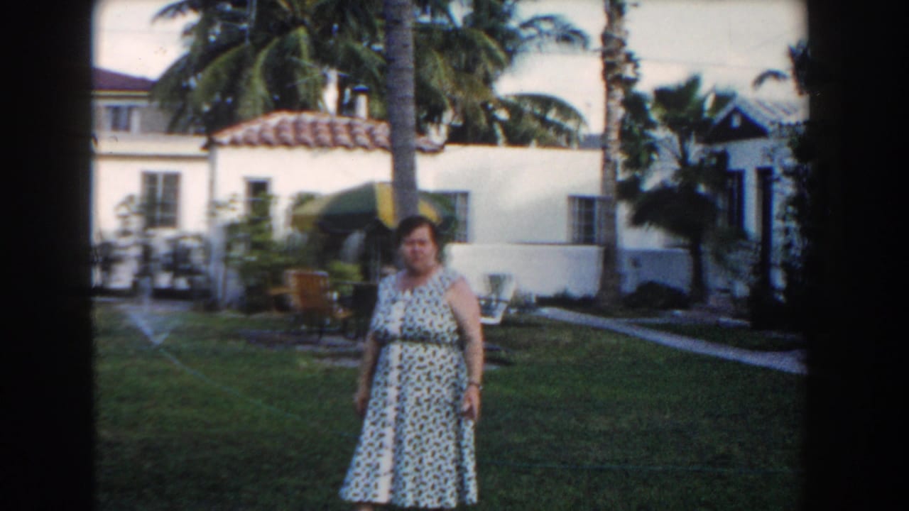 In 1960 Miami home-movie footage, an adult woman stands in a tropical residential garden with palms and a white stucco house behind her.